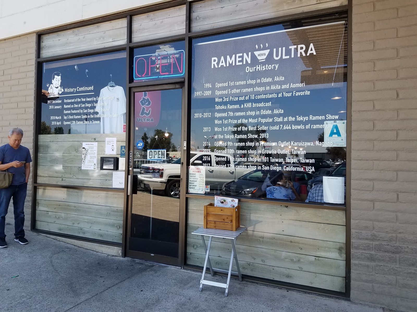 Exterior of Menya Ultra Ramen in Kearny Mesa San Diego with a history wall of awards and an OPEN sign visible through the storefront