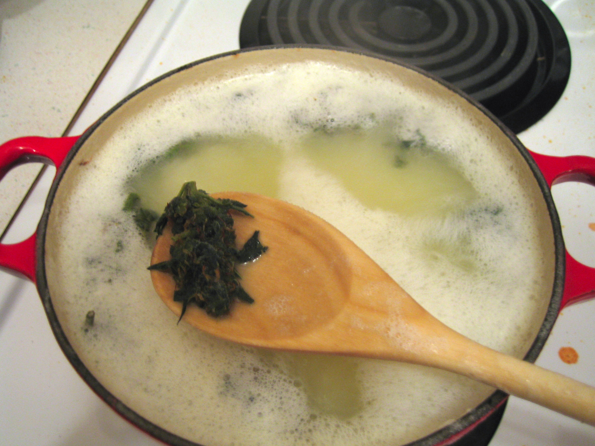 Cannabis flower being stirred into melted butter and water in a red enameled pot on a stove