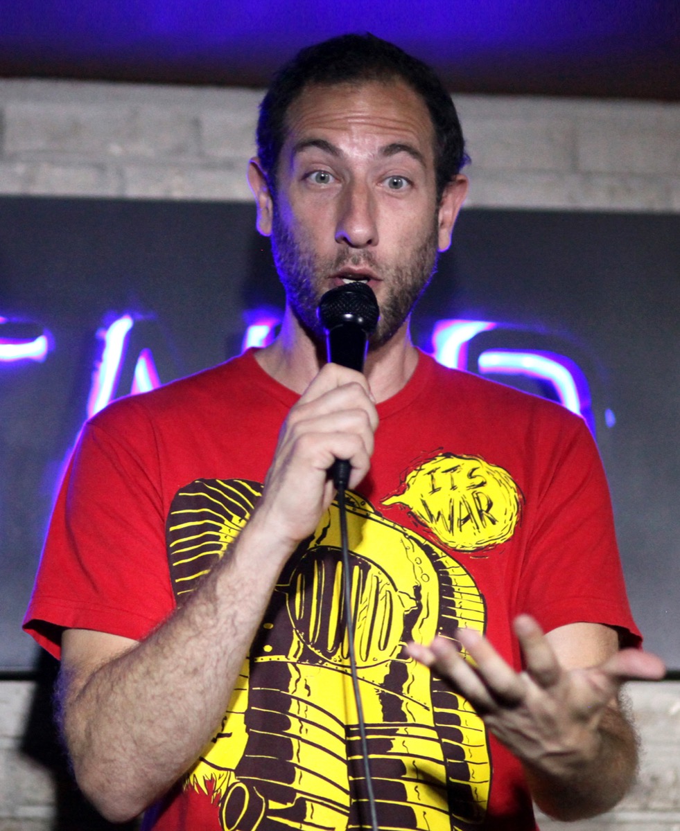 Stand-up comedian Ari Shaffir on stage in a red t-shirt, the loose-cannon long-form cannabis storyteller and Skeptic Tank podcaster.