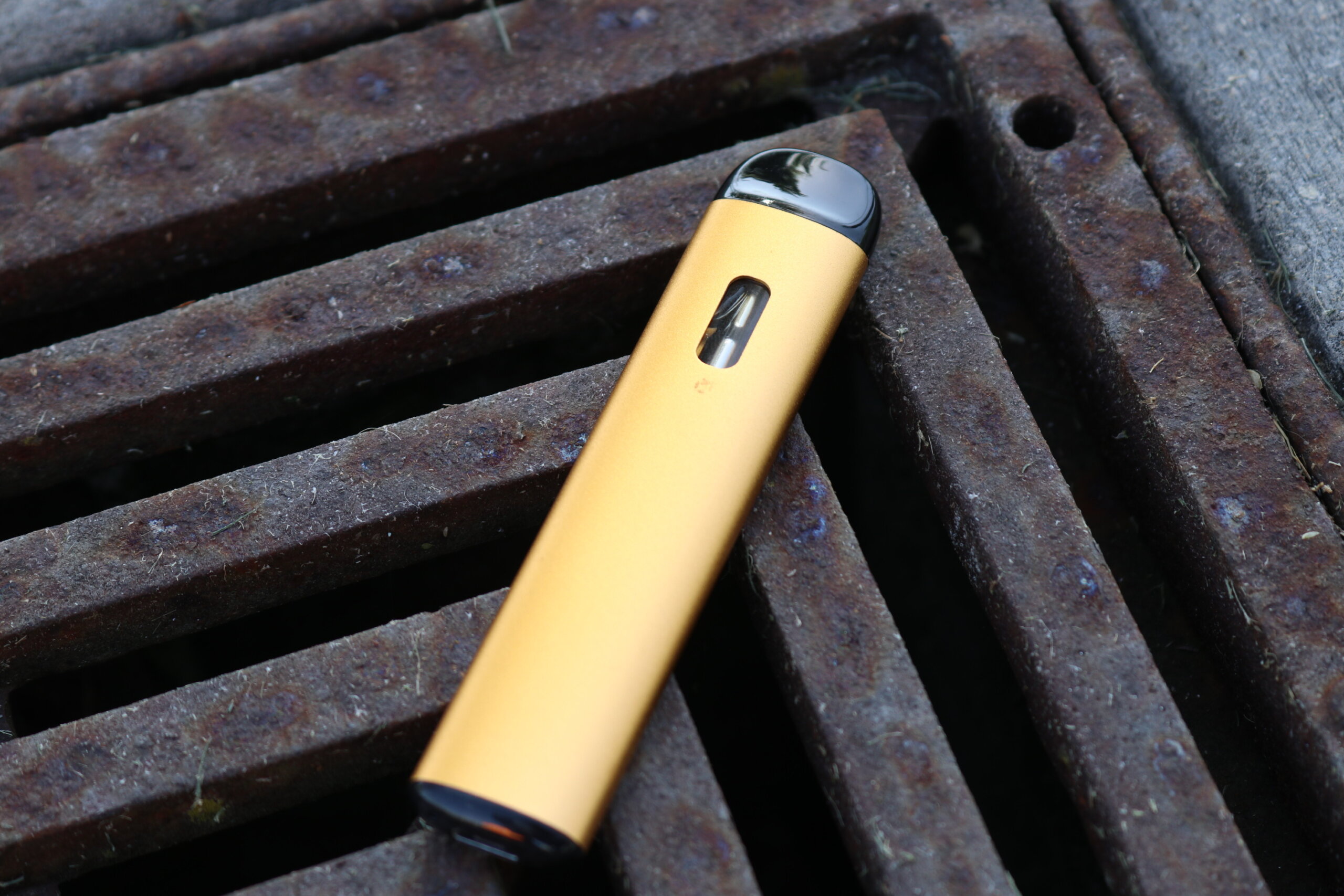 Disposable cannabis vape pen with anodized hardware on a rusted metal grate, representative of the all-in-one vape category Green Thumb Industries distributes under its RYTHM brand across Illinois RISE dispensaries