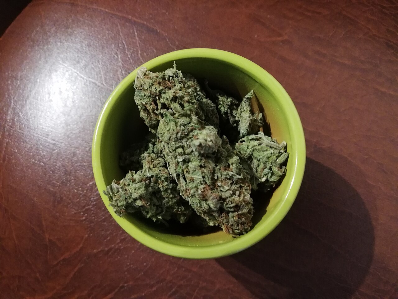 Cannabis flower buds with visible orange pistils and frosty trichomes in a green ceramic dish on a leather counter resembling The Apothecarium Castro flower display