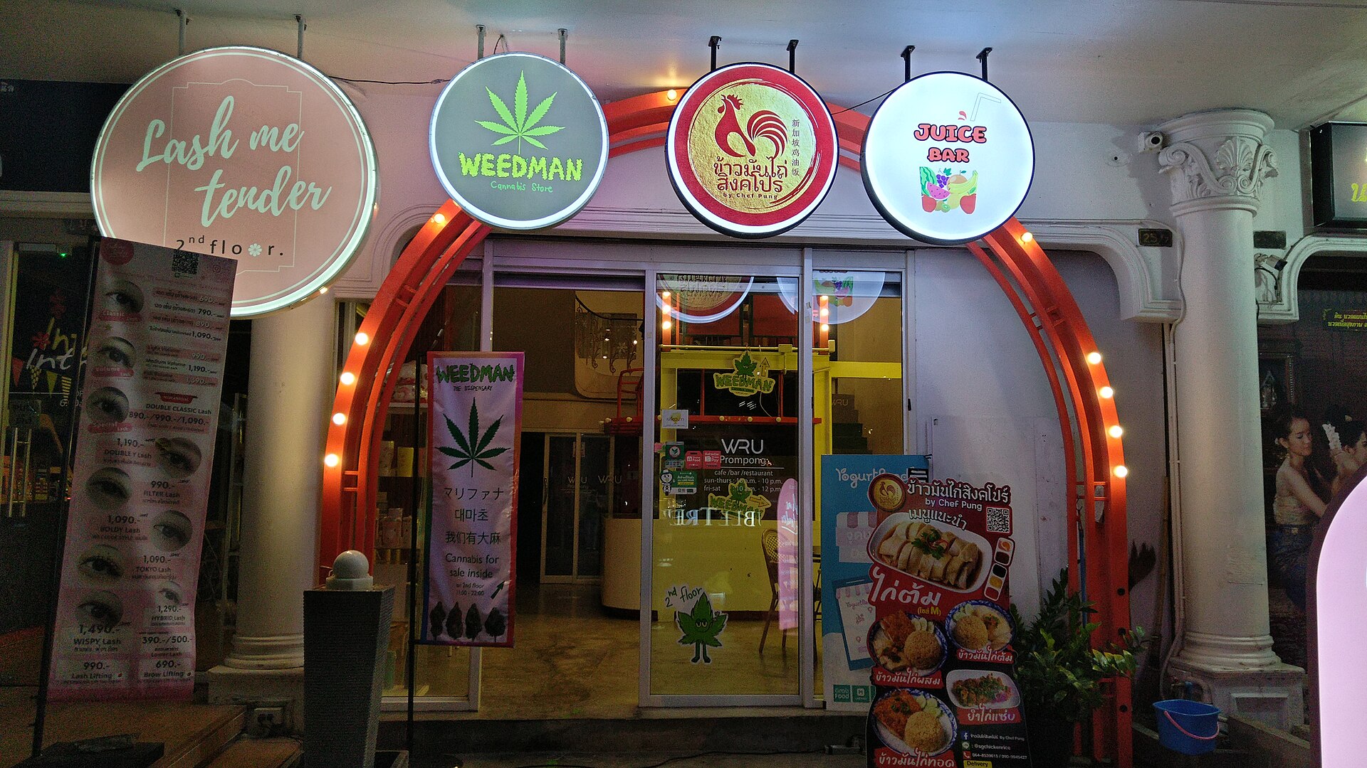 A shopfront in Bangkok with an illuminated Weedman cannabis store sign beside a lash salon and a juice bar
